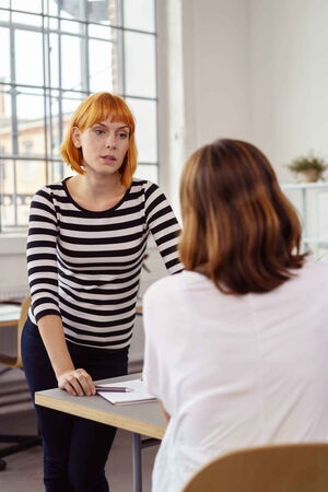 Erfolgreich im Team arbeiten Eine Frau steht vor einer anderen Frau, die mit dem Rücken zu uns sitzt. Die Frau schaut streng. Es scheint einen Konflikt zu geben. Wir helfen Ihnen, Konflikte zu erkennen und zu überwinden und wieder erfolgreich im Team zu arbeiten.